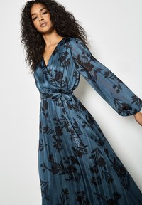 Abito lungo blu con un design avvolgente, maniche lunghe e un motivo floreale in nero. Il tessuto presenta una leggera texture a righe ed è dotato di una cintura da annodare.