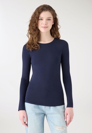 Młoda kobieta o kręconych włosach ubrana w dopasowaną granatową bluzkę z długim rękawem i jasnoniebieskie podarte dżinsy, stojąca na jednolitym tle.