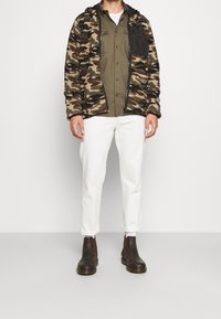 Fleecová bunda se vzorem maskování, černá kapsa na zip, olivově zelená košile, bílé kalhoty a hnědé kotníkové boty.