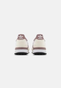 Sneakers with a soft beige suede upper, white fabric accents, and a brown rubber sole. Logo displayed on the heel and tongue in white.