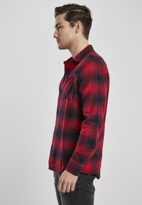Rød og svart rutete flanellskjorte med lange ermer, knappestolpe foran og brystlomme. Stoffet har en teksturert utseende.
