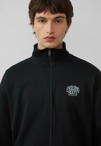 Zwarte ritssportjack gemaakt van zacht materiaal. Voorzien van hoge kraag en geborduurd "EVOLVING ROOTS" logo in lichtblauw.