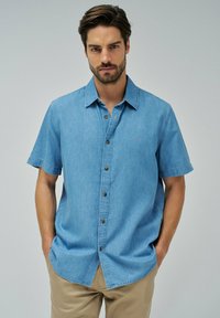 Camisa de mezclilla de manga corta en azul claro con botones en la parte delantera, botones marrones y un pequeño logo bordado en el lado izquierdo del pecho.