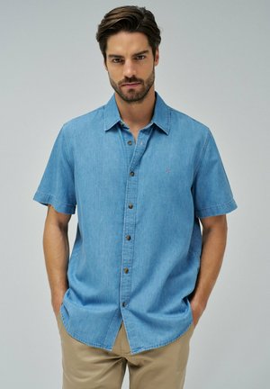 Camisa de mezclilla de manga corta en azul claro con botones en la parte delantera, botones marrones y un pequeño logo bordado en el lado izquierdo del pecho.