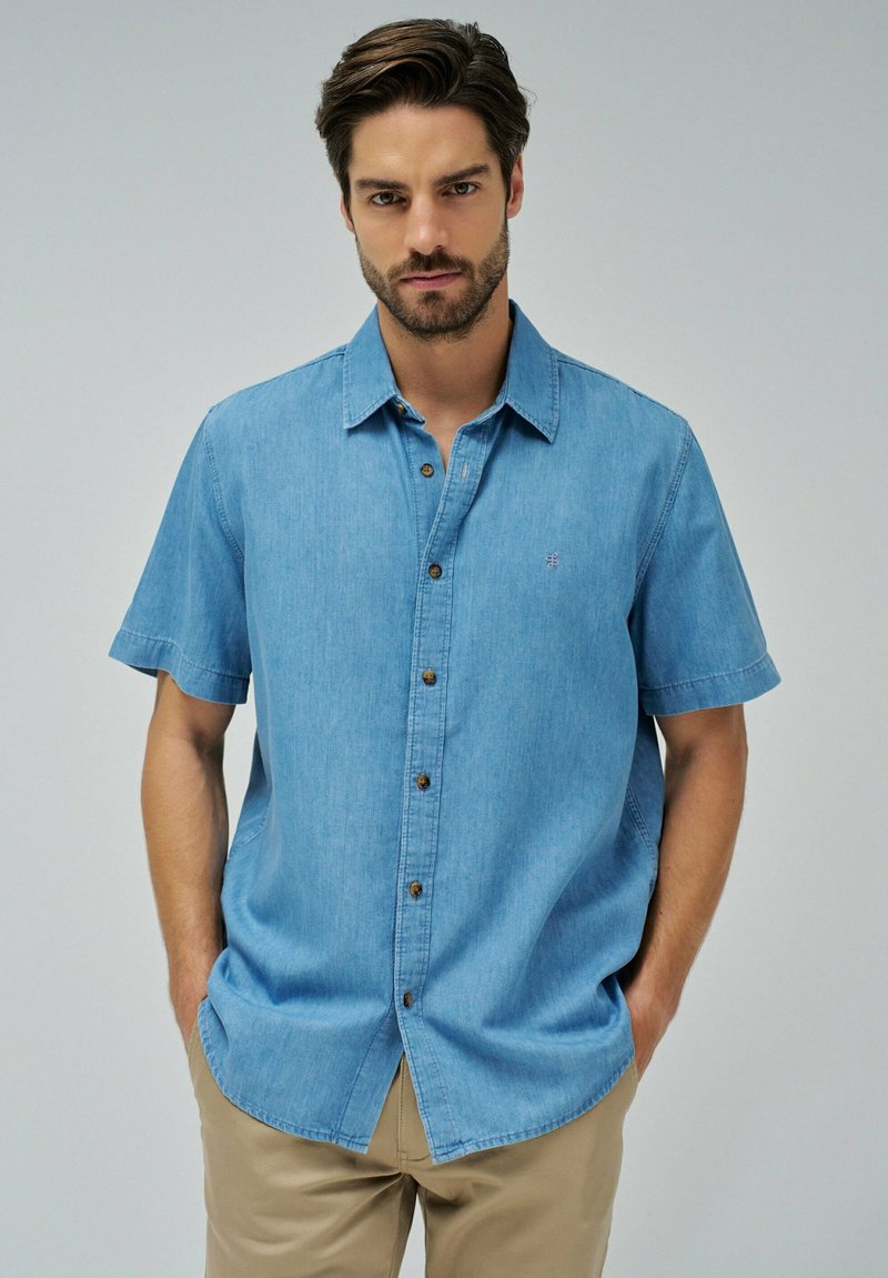 Camisa de mezclilla de manga corta en azul claro con botones en la parte delantera, botones marrones y un pequeño logo bordado en el lado izquierdo del pecho.