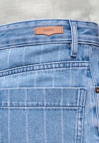 Jean en denim bleu clair avec une grande poche arrière, une passants pour ceinture, et une étiquette de marque en cuir marron portant l'inscription "sessùn" au-dessus de la poche.