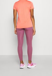Mujer de pie con una blusa corta de manga en color coral, leggings malva y zapatillas deportivas rosas contra un fondo claro y liso.