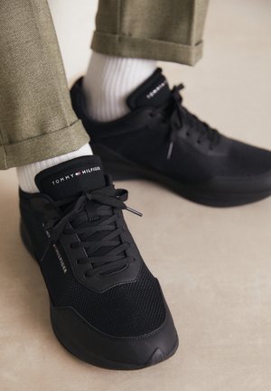 Sorte Tommy Hilfiger sneakers med hvide sokker og beige bukser mod en neutral gulvbaggrund.