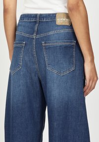 Weite Denim-Jeans in Mittelblau, mit einem strukturierten, hellbraunen Etikett am hinteren Bund, kontrastierenden Nähten und zwei Gesäßtaschen.