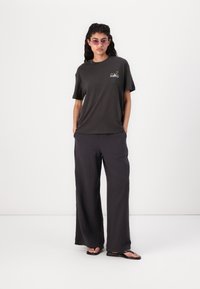 T-shirt grigio scuro con vestibilità comoda, caratterizzato da una piccola grafica sul petto, abbinato a pantaloni ampi e testurizzati grigio scuro e infradito.