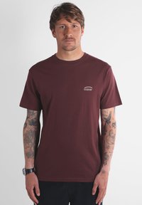 Homme avec des tatouages sur les deux bras portant un t-shirt OXBOW bordeaux et un pantalon noir, se tenant contre un fond blanc uni.