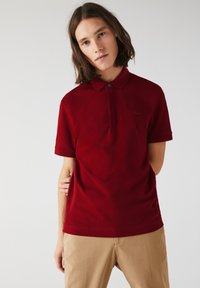 Röd kortärmad pikétröja med krage, som har en diskret broderad logotyp på bröstet och knapplist. Slät tygtextur.