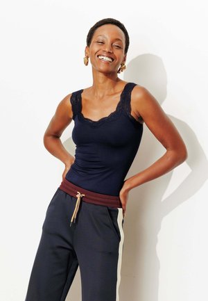 Femme souriante aux cheveux courts portant un débardeur bleu marine orné de dentelle et un pantalon de survêtement bleu marine avec une bande blanche, debout les mains sur les hanches contre un mur blanc.