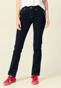 Granatowe dżinsy w wąskim kroju, z kieszeniami z przodu, zapinane na zamek błyskawiczny, z kontrastowymi złotymi przeszyciami, w połączeniu z czerwonymi trampkami.