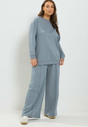 Kvinde står iført løs lyseblå sweatshirt og matchende vidde bukser med hvide sneakers mod en ensfarvet baggrund.