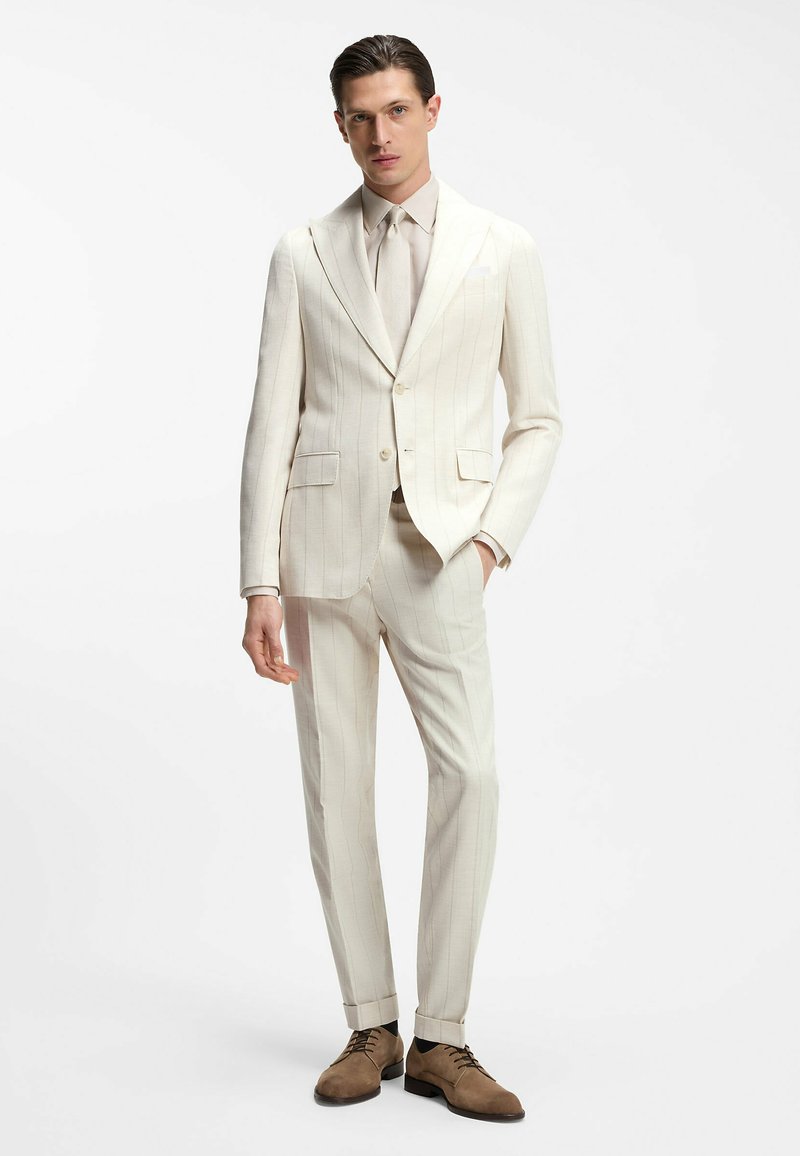 Homme debout portant un costume à fines rayures écru avec une chemise et une cravate assorties, ainsi que des chaussures habillées en daim marron, sur un fond blanc uni.