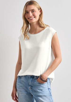 Ung kvinde med blondt hår iført en ærmeløs hvid top og lyseblå jeans, smilende med den ene hånd i lommen.