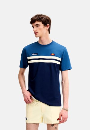 Young man wearing blue and navy striped ellesse t-shirt and beige shorts with small logos, holding glasses, looking left against plain background.