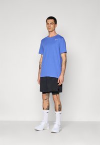 Hombre con tatuajes que lleva una camiseta azul de Nike, pantalones cortos negros en capas, calcetas blancas de Nike y zapatillas blancas, de pie contra un fondo liso.