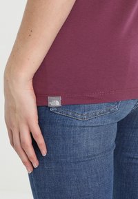 Maroon t-paita, jossa on pehmeä texture ja lyhyet hihat, ja jossa on harmaa merkkilappu helman kohdalla, yhdistettynä sinisiin farkkuihin.