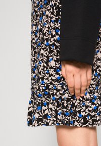 Mano che riposa accanto a una gonna orlata con un motivo floreale nero, beige e blu, indossando una manica lunga nera.