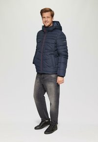 Marineblauwe gewatteerde jas met capuchon, voorzien van een rits aan de voorkant, twee borstzakken, rode accenten in de stiksel, gedragen met grijze jeans en zwarte sneakers.