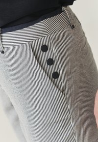 Pruhované kalhoty v námořnické modré a bílé látce s boční kapsou a třemi černými knoflíky. Textura vypadá mírně tkaná s upraveným střihem.