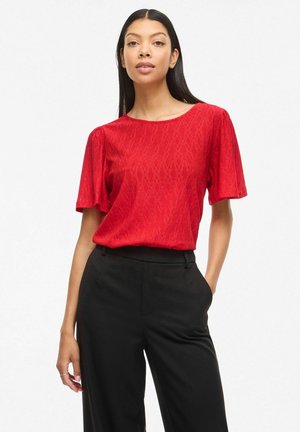 Femme debout, une main dans la poche, portant un chemisier rouge texturé à manches courtes et un pantalon noir taille haute, sur fond blanc.