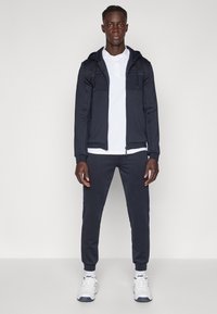 Tumši zils treniņtērps ar rāvējslēdzēja kapuci, divām krūšu kabatām, zem tā balts polo krekls un tumši zilas sporta bikses. Valkāts kopā ar baltām sporta kurpēm.