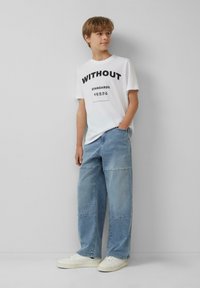 Witte katoenen T-shirt met zwarte tekst, gecombineerd met lichtblauwe broek met wijdbeens en een patchworkontwerp, en witte sneakers met een gestructureerde afwerking.