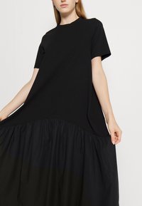 Schwarzes Kleid mit kurzen Ärmeln, einem runden Halsausschnitt und einem voluminösen, gerafften Rock. Hergestellt aus einem glatten Stoff mit weicher Textur.