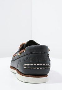 Scarpe da barca in pelle nera con accenti marroni, suola bianca, cuciture visibili e logo inciso sul retro. Presentano occhielli dorati.