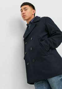 Cappotto doppiopetto blu navy con grandi bottoni, colletto a rever, tessuto misto lana e tasche laterali, abbinato a una camicia di jeans e jeans.