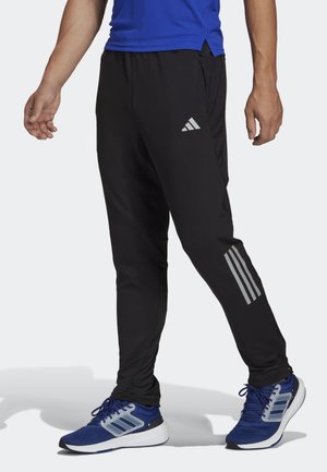 Mężczyzna noszący czarne spodnie dresowe Adidas z odblaskowymi logo oraz niebieskie buty do biegania Adidas.