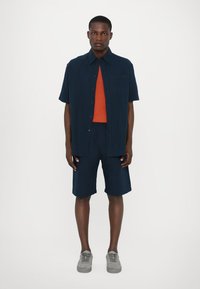 Chemise à manches courtes bleu marine à boutons sur un haut orange, associée à un short bleu marine et des baskets grises. Tissu lisse, coupe décontractée, sans motifs.