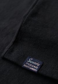 Tissu en coton noir avec une étiquette cousue en bleu portant un texte multicolore indiquant "Superdry" et les mots "Stylé & Fabriqué, Vêtements de Qualité."