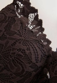 Mørkebrun blonderstof med indviklede blomster mønstre og bølgede kanter. Teksturen fremstår delikat med åbne blondearbejdsdetaljer.