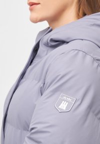 Gepolsterte Jacke in Hellgrau, ausgestattet mit einer Kapuze, vorderen Knöpfen und einem Logoschild mit einem Burg-Design auf dem linken Ärmel. Glatte Textur.