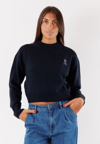 Maglione corto blu navy con scollo e polsini a coste, caratterizzato da un piccolo logo ricamato sul petto. Abbinato a jeans blu a vita alta.
