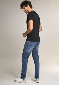 Camiseta negra de manga corta, jeans azules y zapatillas blancas con acentos verdes; mostrado desde la parte de atrás, destacando el ajuste y el estilo casual.