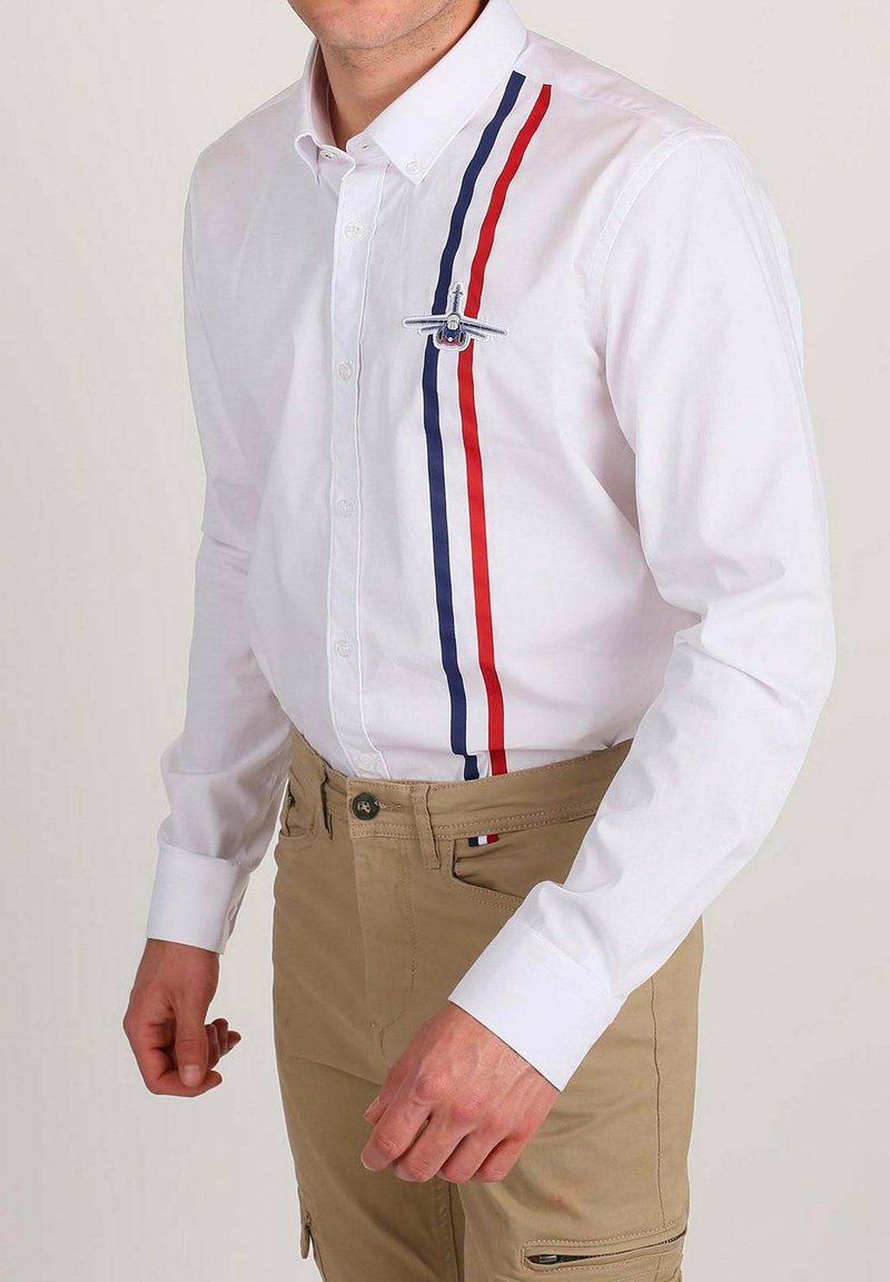 Homme portant une chemise blanche avec des rayures verticales rouges et bleu marine et un écusson d'avion sur la poitrine, accompagnée de pantalons beiges.