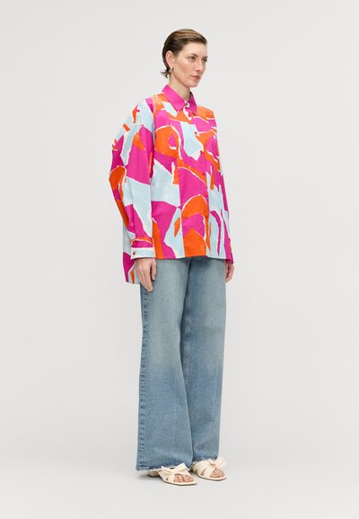 Colorful oversized button-up shirt with pink, blue, and orange abstract patterns; paired with wide-leg blue denim jeans and light sandals.