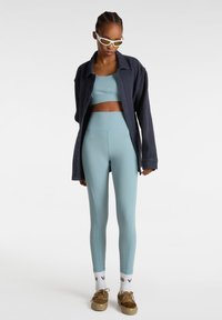 Un reggiseno sportivo azzurro chiaro e leggings a vita alta con cuciture a contrasto, abbinati a una camicia oversized scura con bottoni e scarpe con stampa animale.
