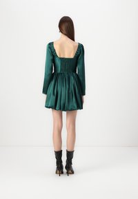 Grön satinklänning med en figursydd bodice, puffärmar och en utsvängd kjol. Har en fyrkantig rygg och smockdetalj. Svarta klackstövlar.