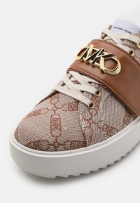 Trampki z brązową tekstylną cholewką ze wzorem, białą gumową podeszwą i brązowym skórzanym paskiem z złotym logo. Białe sznurówki.