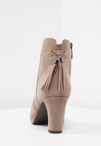 Beige Wildleder-Stiefelette mit einem schmalen hohen Absatz, ausgestattet mit einem Quastenakzent und einem Reißverschluss auf der Rückseite, glatte Textur und schlankes Design.