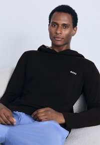 Must kapuutsiga dressipluus, millel on beež sisemine vooder ja väike valge „BOSS" logo. Kandja istub kergelt tekstureeritud pinnal ning kannab helerohelisi pükse.