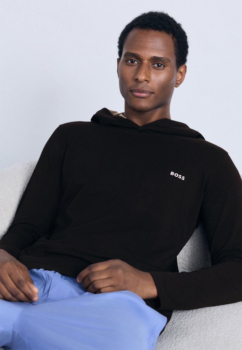 Črna jopa s kapuco z bež notranjo podlogo in majhnim belim logotipom »BOSS«. Oblečena s svetlo modrimi hlačami, sedi na teksturirani površini.