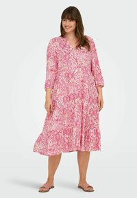 Lungo vestito rosa con motivi floreali e geometrici, scollo a V, maniche a tre quarti e gonna a strati. Indossato con sandali rosa.