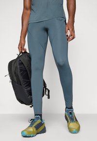 Herre atletikoutfit, der inkluderer teal leggings med en glat tekstur, en tætsiddende trøje og farverige løbesko. Rygsæk i hånden, slank design.